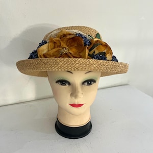 Peut inclure: Chapeau de paille à large bord, orné de grandes fleurs orange séchées, de baies bleues et de feuilles vertes. Le chapeau est présenté sur une tête de mannequin avec du rouge à lèvres rouge et du maquillage foncé. Un chapeau vintage.