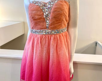 以下が含まれることがあります： ピーチとピンクの色合いの、ストラップレスの膝丈ドレス。ボディスはギャザー入りで、キラキラ光るラインストーンで飾られています。スカートはピーチから鮮やかなピンクへと変化し、グラデーション効果を生み出しています。マネキンに着せられています。