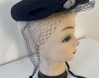 May include: A black vintage hat with a decorative beaded accent and a delicate veil. The hat is displayed on a mannequin head, showcasing its shape and details. The veil drapes gracefully, adding a touch of elegance.