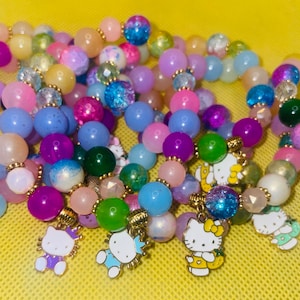 Puede incluir: Una colección de pulseras de cuentas coloridas con una variedad de cuentas de colores pastel, incluyendo rosa, morado, azul y verde. Cada pulsera presenta dijes dorados de un personaje de gato de dibujos animados.
