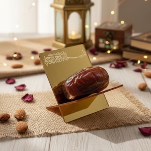 May include: A golden stand with Arabic calligraphy holds a large date. The stand sits on a burlap cloth, surrounded by nuts, rose petals, and a decorative lantern. The scene is lit with warm fairy lights, creating a festive atmosphere.