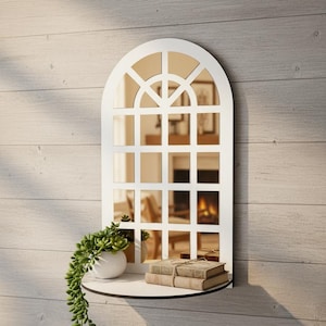 Könnte beinhalten: Weißer, bogenförmiger Fenster-Spiegel mit einem Regal. Der Spiegel hat ein Gittermuster und spiegelt einen Raum mit Kamin wider. Ein kleiner weißer Topf mit einer Sukkulentenpflanze und zwei Büchern befindet sich auf dem Regal. Der Spiegel ist an einer hellen Holzpaneelwand befestigt.