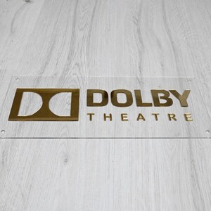 May include: A gold-colored sign with the text "DOLBY THEATRE" on a clear acrylic background.