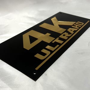 May include: A black rectangular sign with gold lettering that reads "4K ULTRA HD".