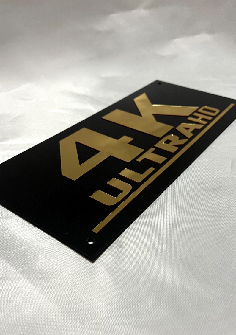May include: A black rectangular sign with gold lettering that reads "4K ULTRA HD".