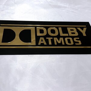 Pode incluir: Uma placa retangular preta com letras douradas que diz "DOLBY ATMOS". O logotipo da Dolby est&aacute; &agrave; esquerda. A placa tem uma borda dourada e &eacute; provavelmente para um sistema de home theater ou entretenimento.