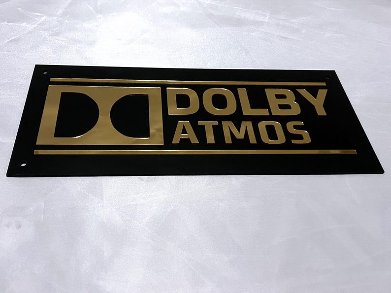Pode incluir: Uma placa retangular preta com letras douradas que diz "DOLBY ATMOS". O logotipo da Dolby est&aacute; &agrave; esquerda. A placa tem uma borda dourada e &eacute; provavelmente para um sistema de home theater ou entretenimento.