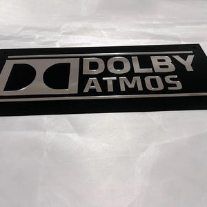Pode incluir: Uma placa retangular preta com as palavras "DOLBY ATMOS" em prata e o logotipo Dolby. O design &eacute; elegante e moderno, provavelmente para equipamentos de &aacute;udio.
