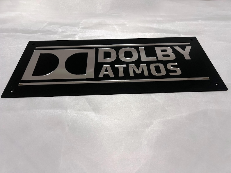 Pode incluir: Uma placa retangular preta com as palavras "DOLBY ATMOS" em prata e o logotipo Dolby. O design &eacute; elegante e moderno, provavelmente para equipamentos de &aacute;udio.