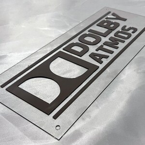 Pode incluir: Uma placa retangular de acr&iacute;lico transparente com as palavras "DOLBY ATMOS" em letras em relevo cinza escuro. O logotipo da Dolby tamb&eacute;m &eacute; cinza escuro. A placa tem cantos arredondados e pequenos orif&iacute;cios.
