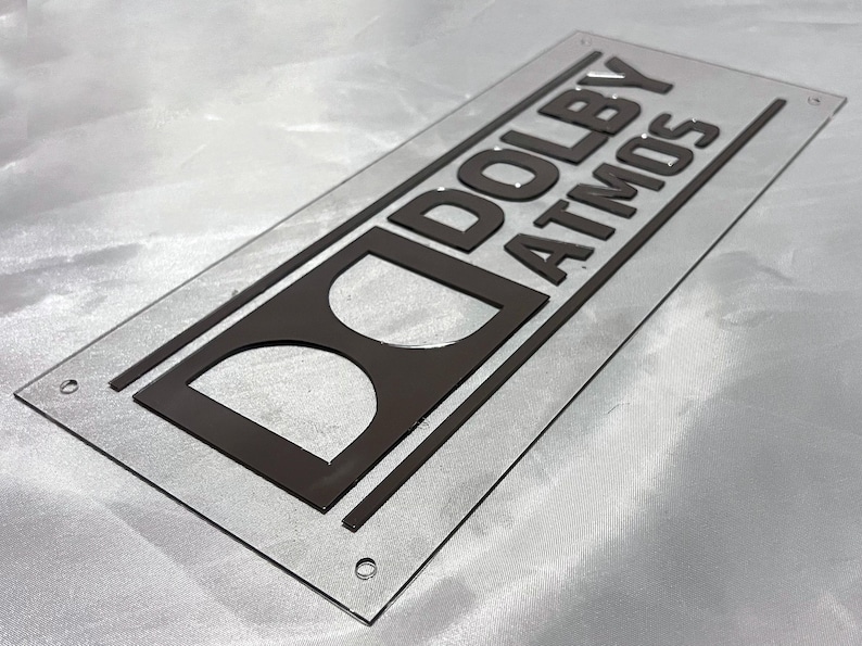 Pode incluir: Uma placa retangular de acr&iacute;lico transparente com as palavras "DOLBY ATMOS" em letras em relevo cinza escuro. O logotipo da Dolby tamb&eacute;m &eacute; cinza escuro. A placa tem cantos arredondados e pequenos orif&iacute;cios.
