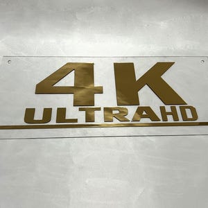 May include: A clear acrylic sign with gold lettering that reads "4K ULTRA HD".