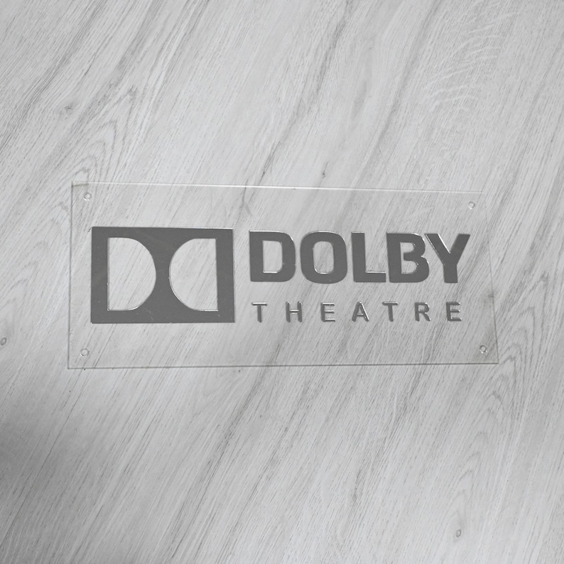 May include: A clear acrylic sign with a silver Dolby Theatre logo. The logo features the Dolby logo and the words "Dolby Theatre".