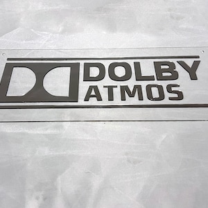 Pode incluir: Um letreiro retangular transparente com as palavras "DOLBY ATMOS" em letras cinza escuro. O logotipo da Dolby tamb&eacute;m &eacute; cinza escuro. O letreiro tem cantos arredondados e pequenos orif&iacute;cios.