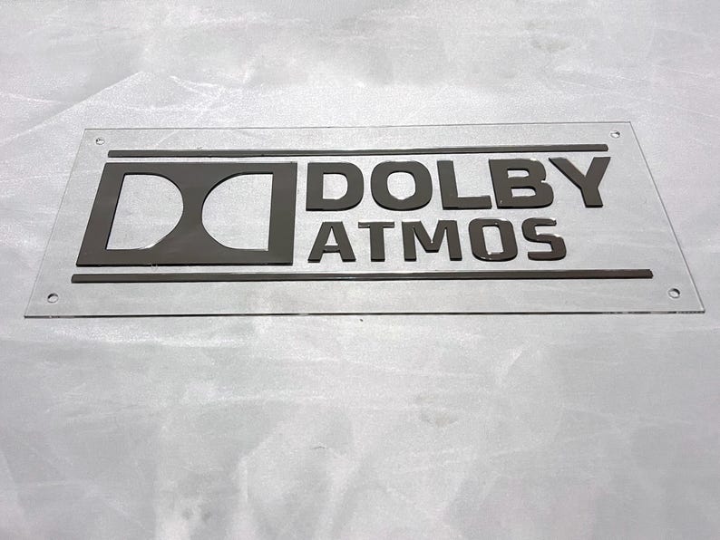 Pode incluir: Um letreiro retangular transparente com as palavras "DOLBY ATMOS" em letras cinza escuro. O logotipo da Dolby tamb&eacute;m &eacute; cinza escuro. O letreiro tem cantos arredondados e pequenos orif&iacute;cios.
