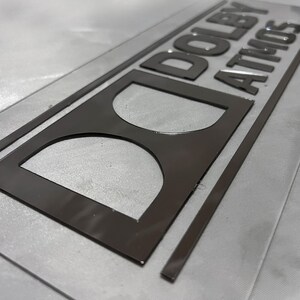Pode incluir: Uma placa retangular com as palavras "DOLBY ATMOS" em letras cinza escuro em relevo. A placa tamb&eacute;m apresenta um logotipo "D" estilizado. A placa est&aacute; em uma superf&iacute;cie cinza claro.