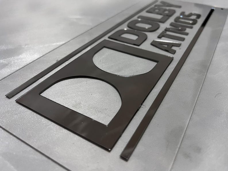 Pode incluir: Uma placa retangular com as palavras "DOLBY ATMOS" em letras cinza escuro em relevo. A placa tamb&eacute;m apresenta um logotipo "D" estilizado. A placa est&aacute; em uma superf&iacute;cie cinza claro.