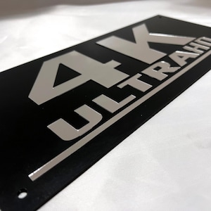 May include: Black rectangular sign with silver lettering that reads "4K ULTRA HD".