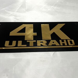 May include: A black rectangular sign with gold lettering that reads "4K ULTRA HD".
