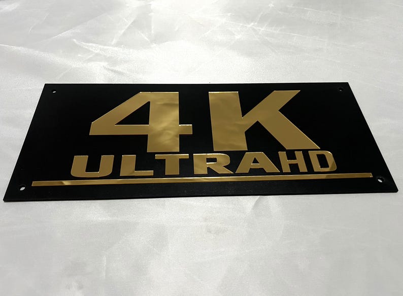 May include: A black rectangular sign with gold lettering that reads "4K ULTRA HD".