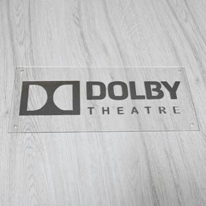 May include: A clear acrylic sign with black lettering that reads "Dolby Theatre". The Dolby logo is a stylized "D" with a circle inside.