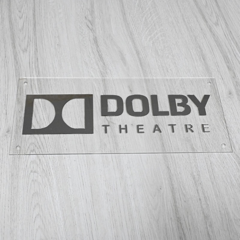 May include: A clear acrylic sign with black lettering that reads "Dolby Theatre". The Dolby logo is a stylized "D" with a circle inside.