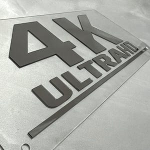 May include: A clear acrylic sign with the text "4K ULTRA HD" in black raised lettering.