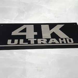 May include: A black rectangular sign with silver lettering that reads "4K ULTRA HD".