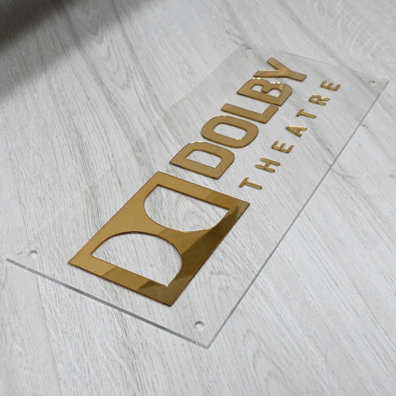 May include: A clear acrylic sign with a gold-colored "Dolby Theatre" logo. The logo features a stylized "D" with two circles inside.