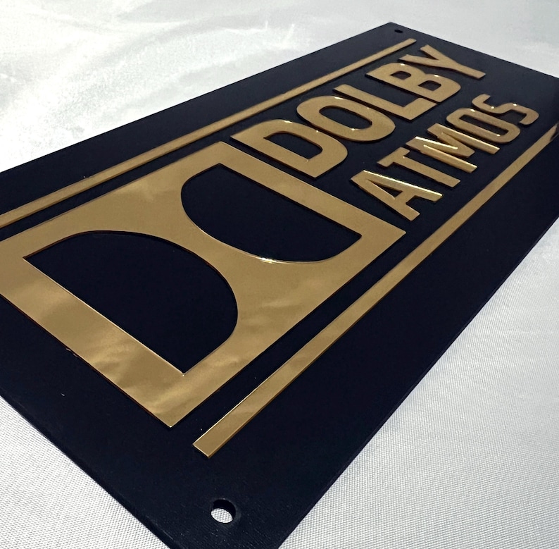 Pode incluir: Uma placa retangular preta com o texto dourado "DOLBY ATMOS" e o logotipo da Dolby. As letras e o logotipo dourados s&atilde;o em relevo. A placa est&aacute; angulada, mostrando as letras douradas.
