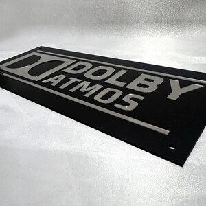 Pode incluir: Uma placa retangular preta com a inscri&ccedil;&atilde;o prateada "DOLBY ATMOS". A placa tem um design elegante e moderno, provavelmente para home theater ou equipamentos de &aacute;udio.