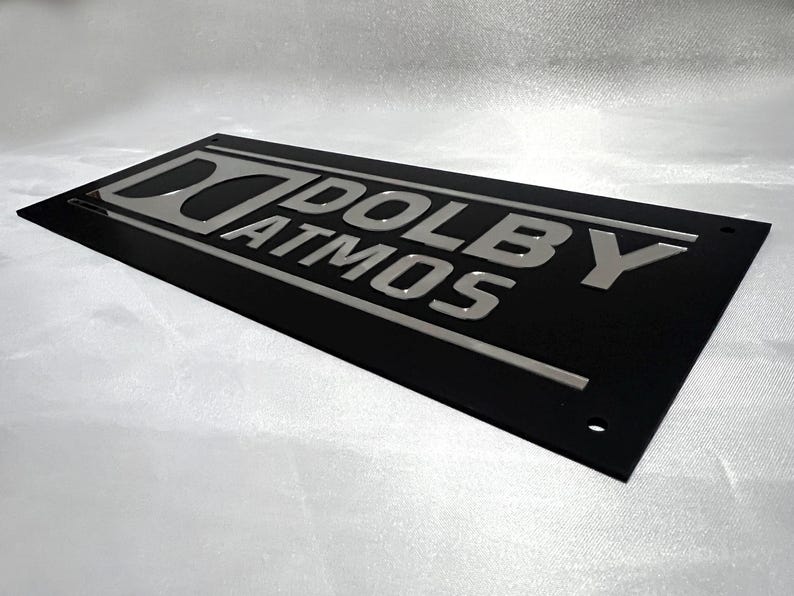 Pode incluir: Uma placa retangular preta com a inscri&ccedil;&atilde;o prateada "DOLBY ATMOS". A placa tem um design elegante e moderno, provavelmente para home theater ou equipamentos de &aacute;udio.