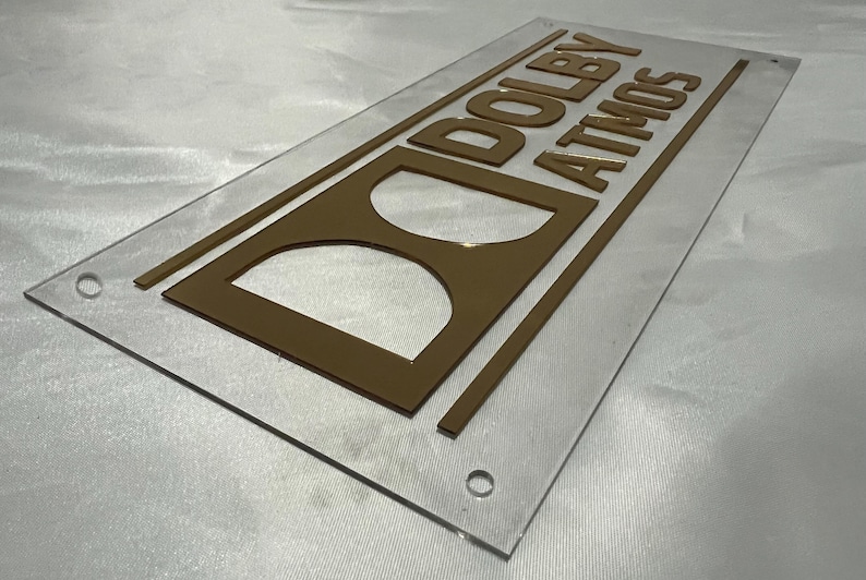 Pode incluir: Uma placa de acr&iacute;lico transparente com as palavras "DOLBY ATMOS" em letras douradas. A placa apresenta o logotipo da Dolby e &eacute; retangular com cantos arredondados. Existem orif&iacute;cios nos cantos para montagem.