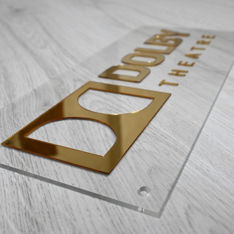 May include: A clear acrylic sign with gold lettering that reads "DOLBY THEATRE". The sign has a gold cutout of the Dolby logo.