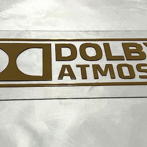 Pode incluir: Uma placa retangular transparente com as palavras "DOLBY ATMOS" em letras douradas. A placa tem um logotipo Dolby dourado e duas linhas horizontais douradas. Existem quatro pequenos orif&iacute;cios nos cantos da placa.