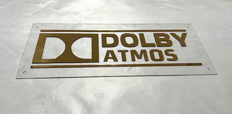 Pode incluir: Uma placa retangular transparente com as palavras "DOLBY ATMOS" em letras douradas. A placa tem um logotipo Dolby dourado e duas linhas horizontais douradas. Existem quatro pequenos orif&iacute;cios nos cantos da placa.