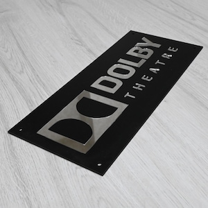 May include: Black sign with a silver "Dolby Theatre" logo. The "Dolby" text is in a bold, sans-serif font, and the "Theatre" text is in a smaller, sans-serif font.