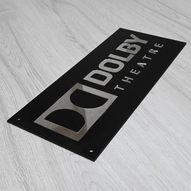 May include: Black sign with a silver "Dolby Theatre" logo. The "Dolby" text is in a bold, sans-serif font, and the "Theatre" text is in a smaller, sans-serif font.