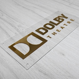 May include: A clear acrylic sign with gold lettering that reads "Dolby Theatre".