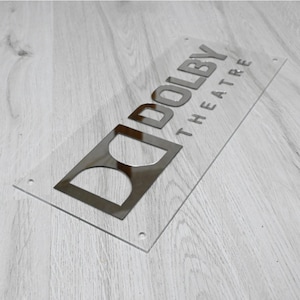 May include: A clear acrylic sign with a silver metallic finish. The sign reads "Dolby Theatre".