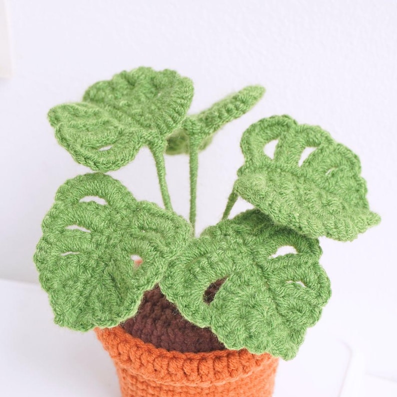 May include: A crocheted green monstera plant in a small orange pot. The plant has several leaves with cutouts.