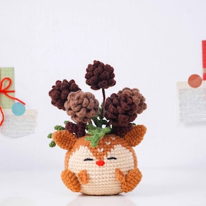 May include: A handmade crochet deer planter with a cream-colored body, orange antlers and ears, and a red nose. The deer holds a cluster of brown, tan, and green crocheted flowers. The planter is a whimsical, decorative item.