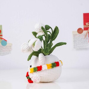 May include: A handmade crochet bunny planter with white snowdrop flowers. The white bunny planter has a colorful scarf around its neck. The snowdrop flowers have green stems and leaves. The bunny planter is a unique home decor item.