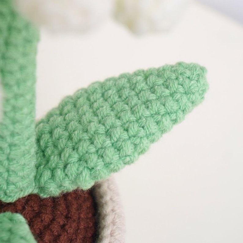 Puede incluir: Primer plano de una planta de crochet verde con una maceta marr&oacute;n y blanca. La planta tiene una superficie texturizada de crochet.