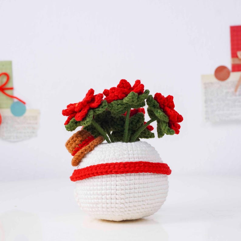 May include: A crocheted poinsettia plant in a white pot with a red stripe. The plant features vibrant red flowers and green leaves. A small brown hat rests on the edge of the pot. The handmade decor is set against a white background.