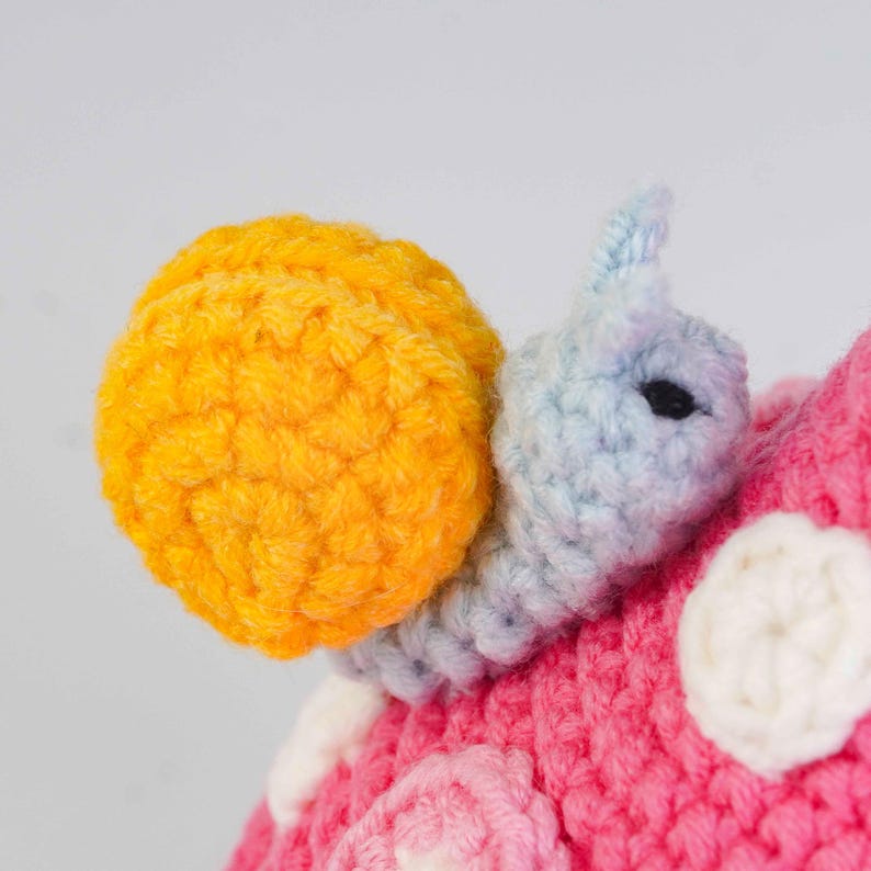 May include: A handmade crochet snail with a bright yellow shell and a blue and gray body. The snail is attached to a pink crocheted surface with white and pink accents. The snail's eye is a small black bead.