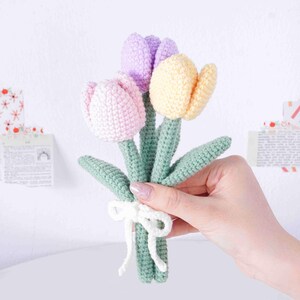 May include: A handmade bouquet of three crocheted tulips in pink, lilac, and yellow. The flowers have green stems and leaves, tied with a white ribbon. The flowers are displayed against a white background.