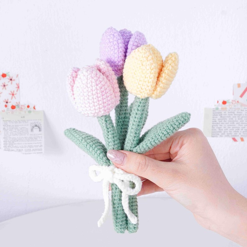 May include: A handmade bouquet of three crocheted tulips in pink, lilac, and yellow. The flowers have green stems and leaves, tied with a white ribbon. The flowers are displayed against a white background.