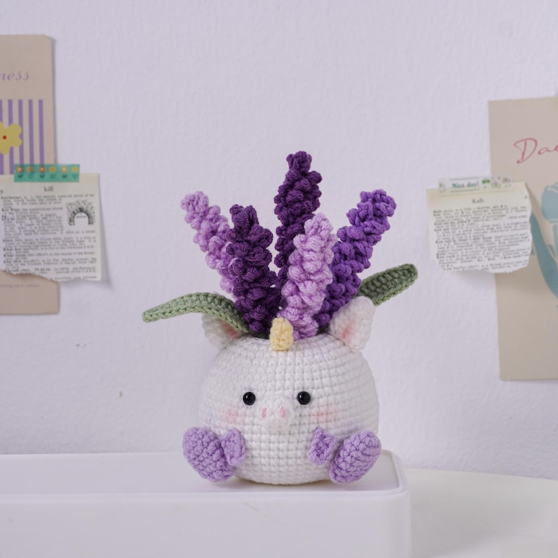 May include: A white crocheted unicorn planter with purple and lavender flowers. The unicorn has a yellow horn, pink cheeks, and purple feet. Green leaves peek out from behind the flowers. A whimsical, handmade home decor item.