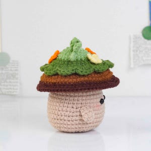 May include: A crocheted mushroom figure with a light brown body, a brown and orange cap, and a green top with orange and yellow accents. The mushroom has small black eyes and rosy cheeks, giving it a friendly appearance.